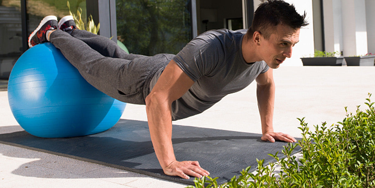 Man doing push-ups on a mat with a blue exercise ball beside him outdoors.